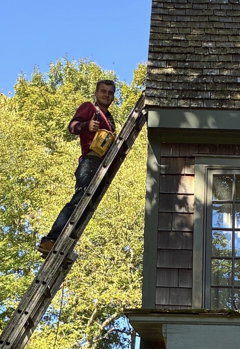 Contractor on a ladder doing exterior shingle and trim work on a colonial-style home