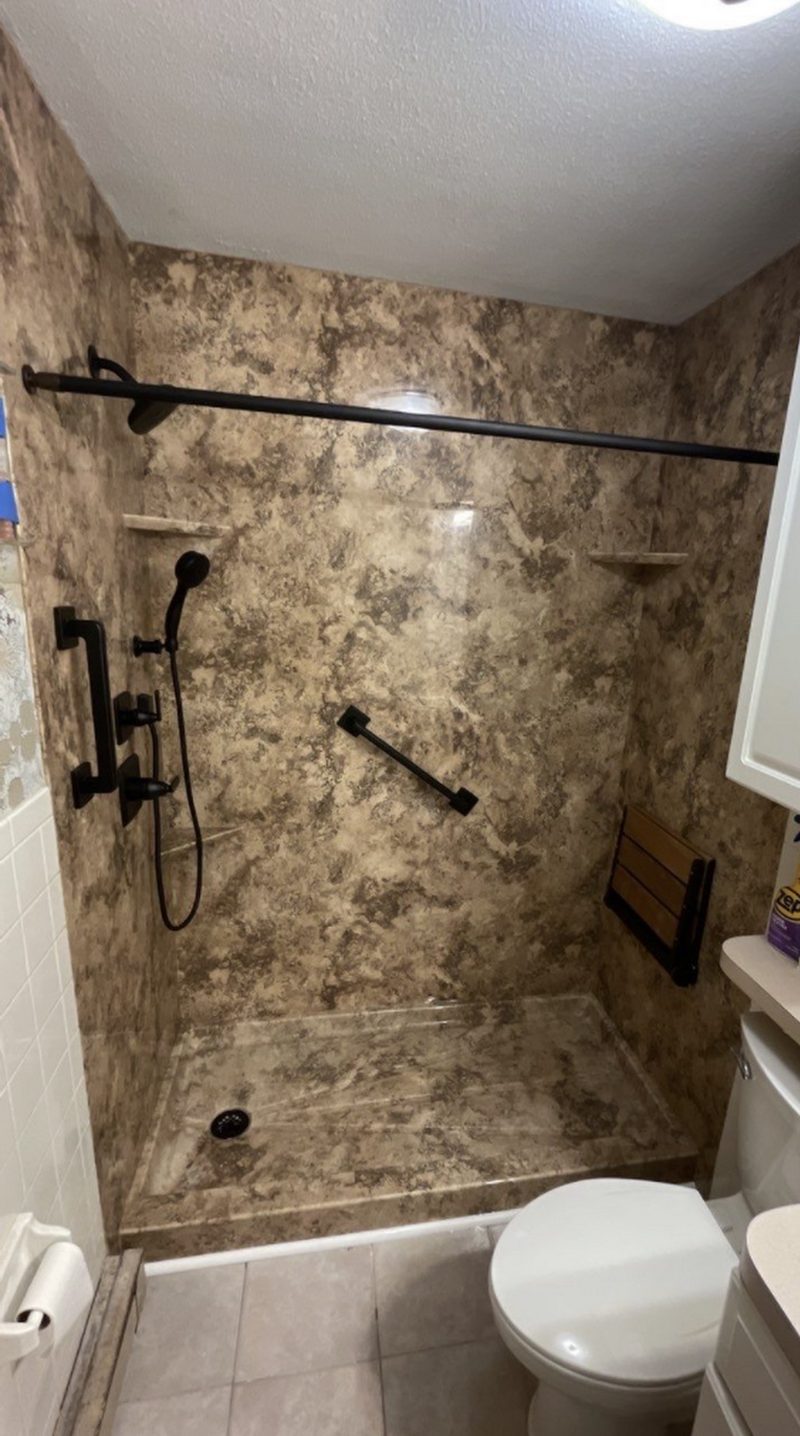 Remodeled shower with one-piece stone-look surround, black fixtures, grab bars and fold-down seat